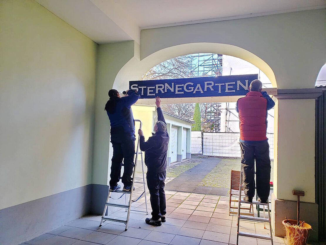 Drei Männer beim Befestigen des Schildes mit der Aufschrift "Sternegarten" in einem Torbogen