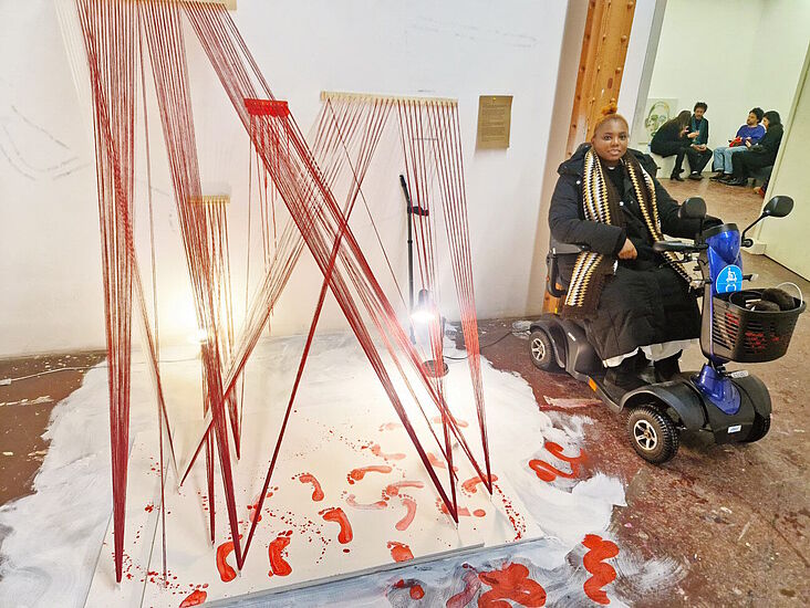 Lillian Munuo und ihr Kunstwerk, welches unter anderem aus vielen roten Stricken besteht; am Boden rote Fußspuren