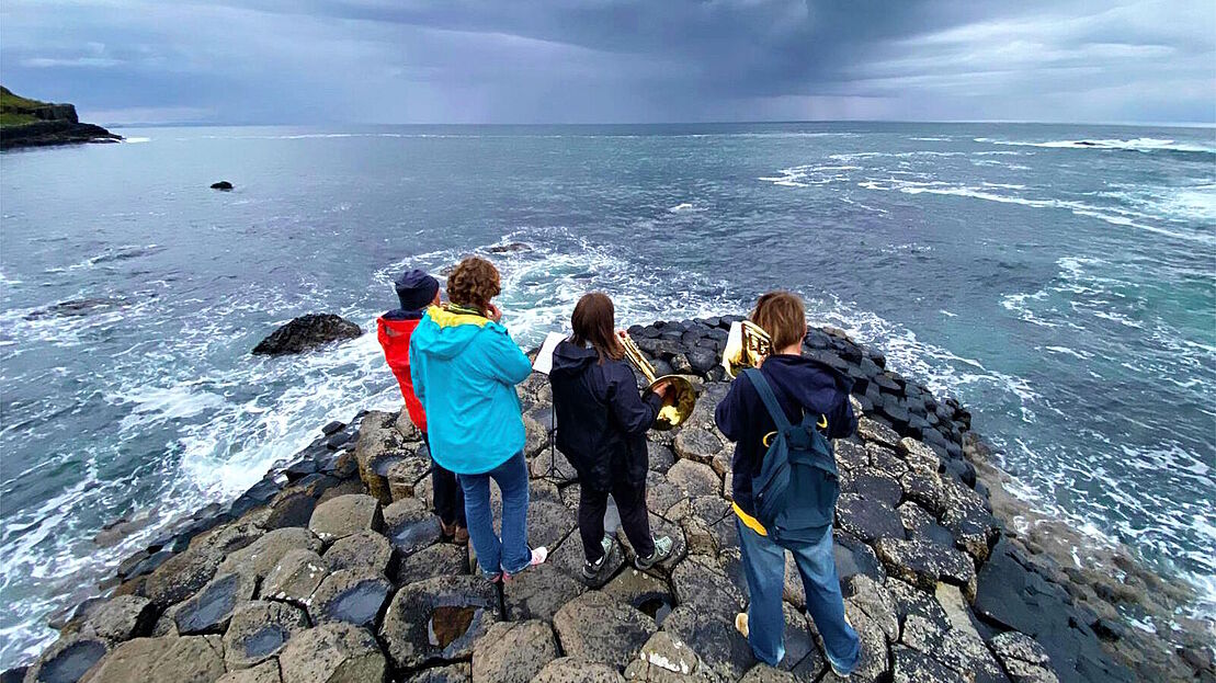 Vier Bläser sind von hinten zu sehen; sie stehen auf Felsen und blasen mit Blick aufs Meer.