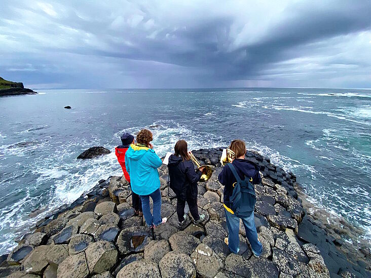 Vier Bläser sind von hinten zu sehen; sie stehen auf Felsen und blasen mit Blick aufs Meer.
