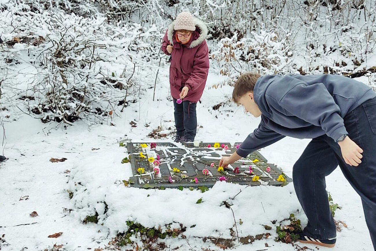 Ein Schüler und eine ältere Frau legen Blumen auf Gedenkstein nieder.