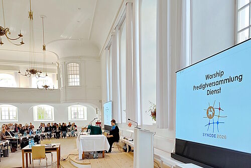 Blick auf Rednerpult und Liturgustisch, im Vordergrund ein Display mit Synoden-Logo