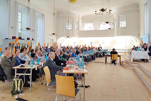 Delegierte im Plenum der Synode, welche ihr orangefarbenen Abstimmungskärtchen hochhalten.