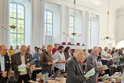 Teilnehmende der Synode stehen an ihren Plätzen im Kirchensaal und singen
