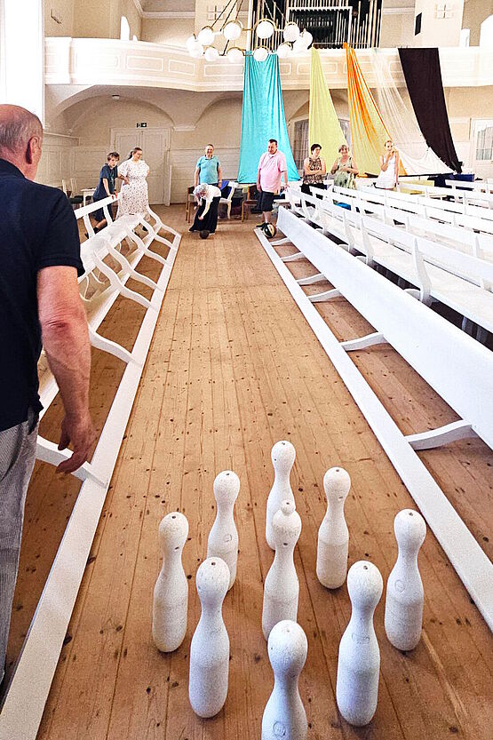 eine improvisierte Kegelbahn im Kirchensaal, die Banden bestehen aus gekippten Saalbänken, im Vordergrund stehen die Pins