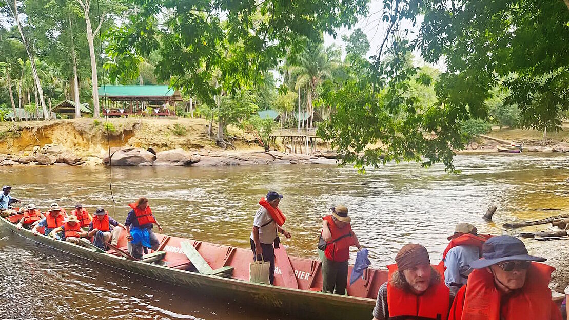 Mehrere Menschen mit Rettungsweste verlassen ein Boot, das am Ufer eines surinamischen Flusses liegt.