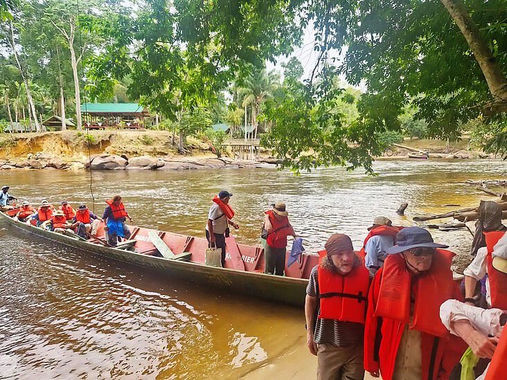 Mehrere Menschen mit Rettungsweste verlassen ein Boot, das am Ufer eines surinamischen Flusses liegt.