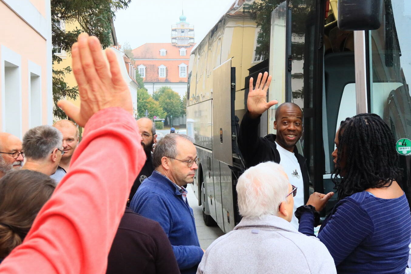 Ein Mann steigt in den Bus und winkt den umstehenden Menschen, im Vordergrund ein winkender Arm
