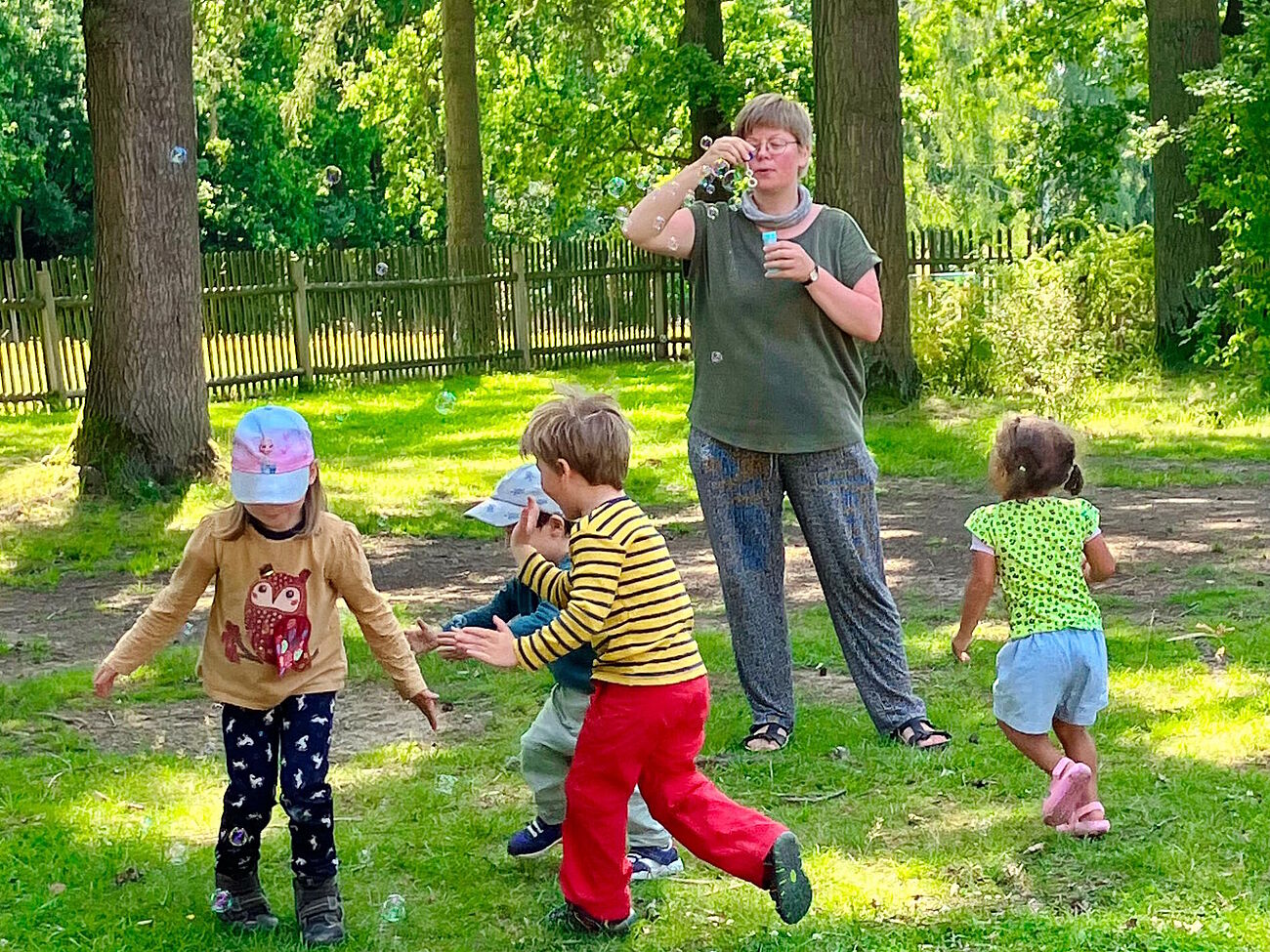 erwachsene Person macht Seifenblasen, Kinder laufen über die Wiese und fangen diese