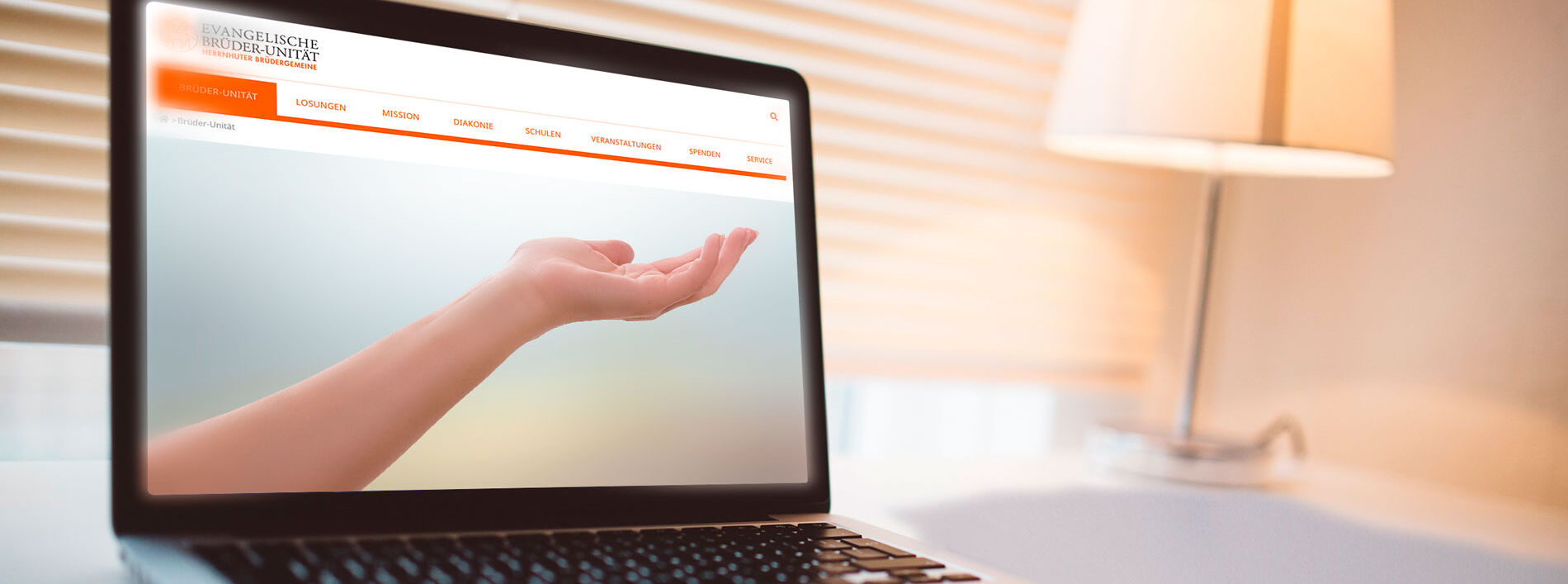 Ein in angenehem warmes Licht getauchter Raum mit einem Laptop im Vordergrund, auf welchem eine Hand ausgestrckte Hand zu sehen ist; im Hintergrund eine Tischlampe, die ein warmes Licht ausstrahlt.