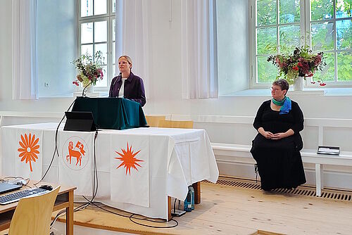Claudia Kusch steht am Liturgustisch und übermittelt ein Grußwort, Erdmuthe D. Frank sitzt daneben auf einer Kirchenbank