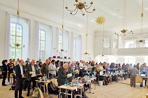 Teilnehmende der Synode stehen an ihren Plätzen im Kirchensaal und singen