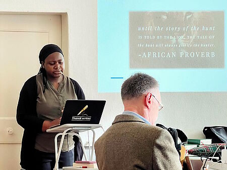 Thandi Soko-de Jong steht vor ihrem Notebook und hält einen Vortrag, im Hintergrund an der Wand ist eine Powerpoint-Präsentation zu sehen