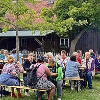 Grillen mit den Mietern Mieterinnen und Mieter beim Grillen zusammensitzend an Biertischgarnituren auf eine Wiese hinter einem Haus.