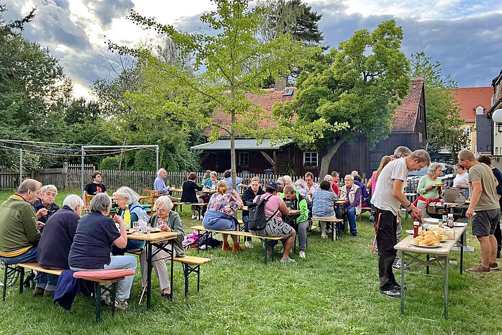 Mieterinnen und Mieter beim Grillen zusammensitzend an Biertischgarnituren auf eine Wiese hinter einem Haus.