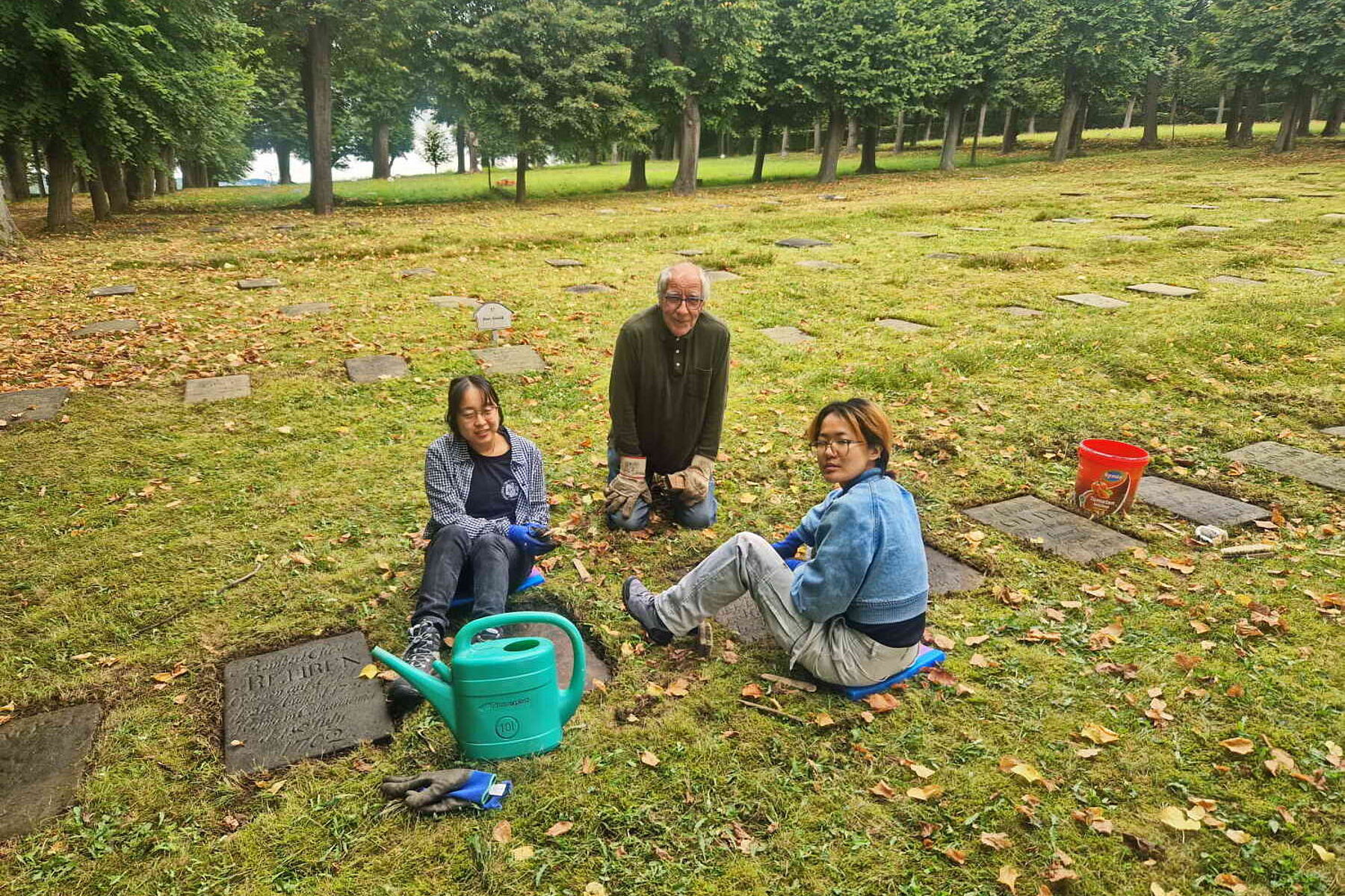 Teilnehmerinnen und ein Gemeindemitglied sitzen auf der Wiese und pausieren von der Reinigung der Grabsteine